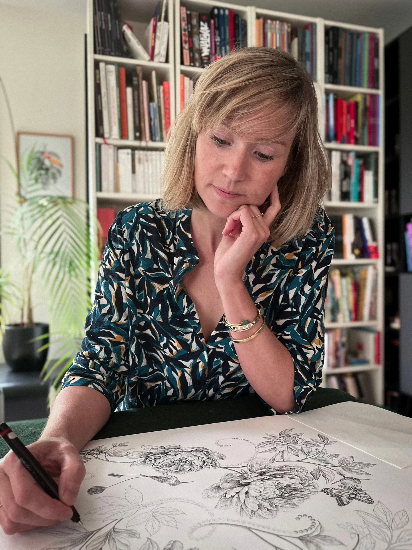 A person sits at a desk beside a large floral drawing, with bookshelves behind.